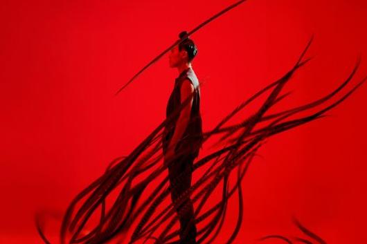 A profile view of a man standing against a red background with black tendrils coming up from the floor.